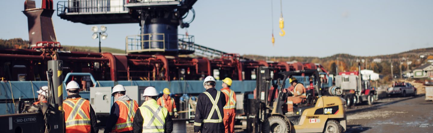 Construction workers in safety gear and high-visibility vests walk across a busy industrial site, with large machinery and equipment visible in the background under clear skies.