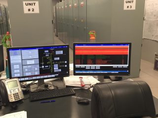 A control room with computer monitors displaying data, a telephone, and various equipment on a desk. In the background are large electrical cabinets labeled 