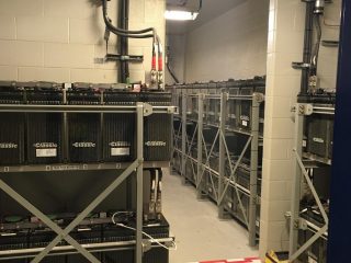 A room with multiple large industrial batteries on metal racks, electrical wiring overhead, and a sign on the floor reading 