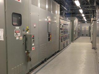 A corridor lined with large industrial electrical panels and control cabinets, labeled with warning signs, inside a facility. The words 