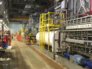 Industrial machine hall with large tanks, pipes, and machinery along the walls; yellow safety railings and various equipment visible, with a wide aisle down the center. Red text reads 