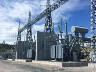 A power station with large transformers, high voltage power lines, and metal structures under a partly cloudy sky. The words 