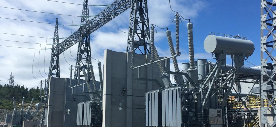 A power station with large transformers, high voltage power lines, and metal structures under a partly cloudy sky. The words "STATION TRANSFORMERS" appear in bold red text at the bottom of the image.