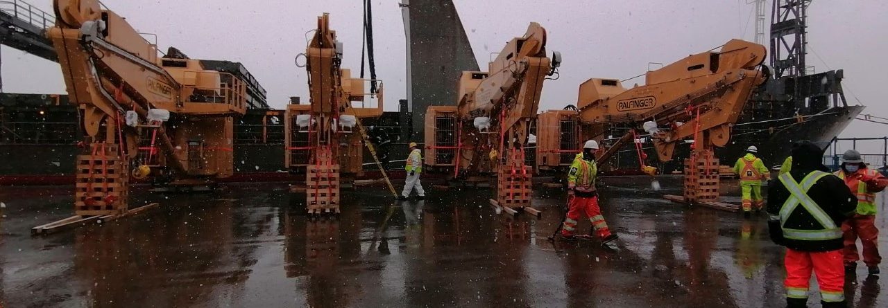 Several workers in safety gear stand on a wet industrial dock in light snowfall, surrounded by large tan cranes and heavy equipment, with a ship and overcast sky in the background.