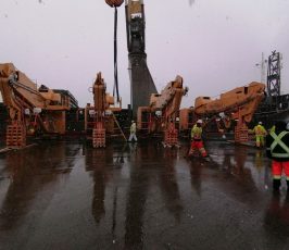 Several workers in safety gear stand on a wet industrial dock in light snowfall, surrounded by large tan cranes and heavy equipment, with a ship and overcast sky in the background.