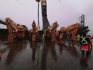 Several workers in safety gear stand on a wet industrial dock in light snowfall, surrounded by large tan cranes and heavy equipment, with a ship and overcast sky in the background.