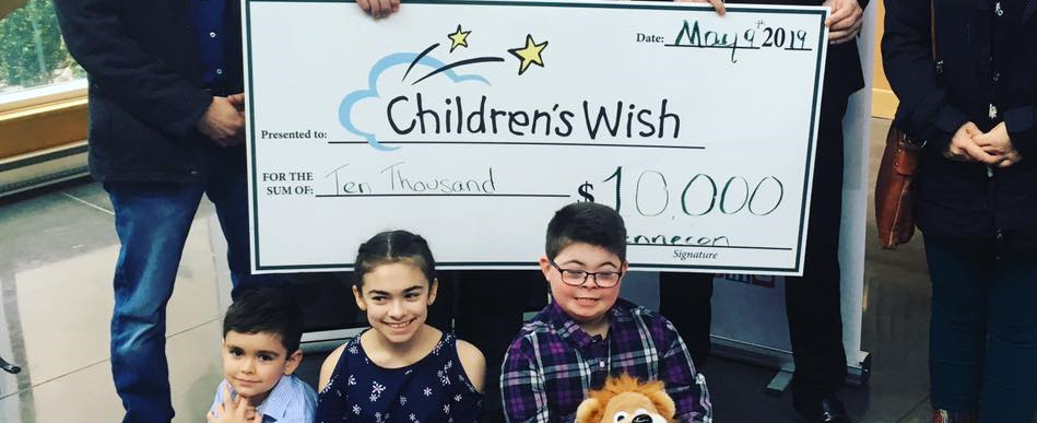 Three children sitting and smiling, with one holding a stuffed animal, in front of a large ceremonial check for $10,000 made out to "Children