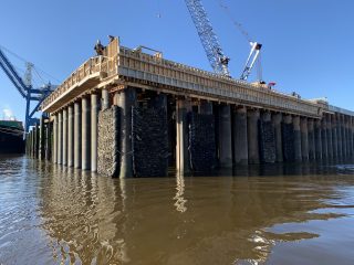 A construction site on the water features a pier supported by large cylindrical pillars; workers and cranes are visible on the wooden deck under a clear blue sky.