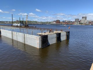 Two workers stand on a large concrete structure floating in a river, with construction equipment and a barge nearby. A city skyline and blue sky with clouds are visible in the background.