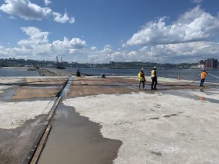 Four construction workers in safety gear work on a dock by the water, handling hoses and equipment on a large concrete and metal surface under a blue sky with scattered clouds.