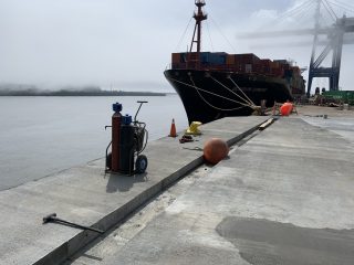 A large cargo ship docked at a foggy port, with containers stacked on deck. Equipment, safety cones, and orange buoys are scattered on the concrete pier. Cranes and misty water are visible in the background.