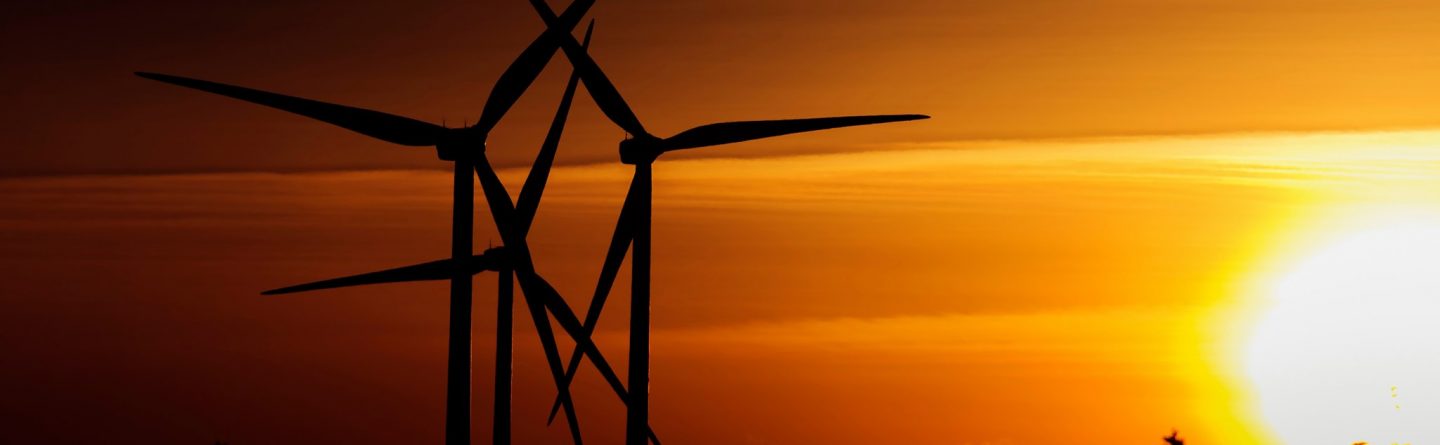 Three wind turbines are silhouetted against a vivid orange sunset sky, with the sun low on the horizon and trees visible in the foreground.