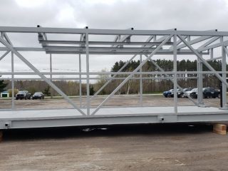 A large metal steel frame structure is being transported on a forklift outdoors, with wooden blocks supporting it on the ground; cars and trees are visible in the background under a cloudy sky.