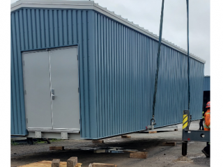 A large blue metal building is being lifted by a crane. Two workers in safety gear stand nearby on a construction site, overseeing the process. Wooden pallets and concrete blocks are on the ground below.