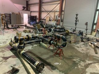 Several large black metal pipes with valves and fittings are assembled on red stands in an industrial workshop, with tools, equipment, and shelves visible in the background.