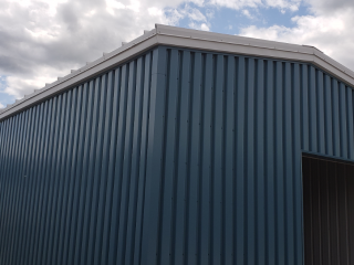 A blue metal building with vertical corrugated panels and a white trim is shown under a cloudy sky. The structure has a large open entryway and no visible windows.