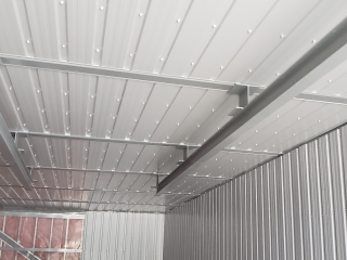 Interior view of a building with corrugated metal walls and ceiling, featuring exposed steel beams and framework, with a partially visible wall in the background.