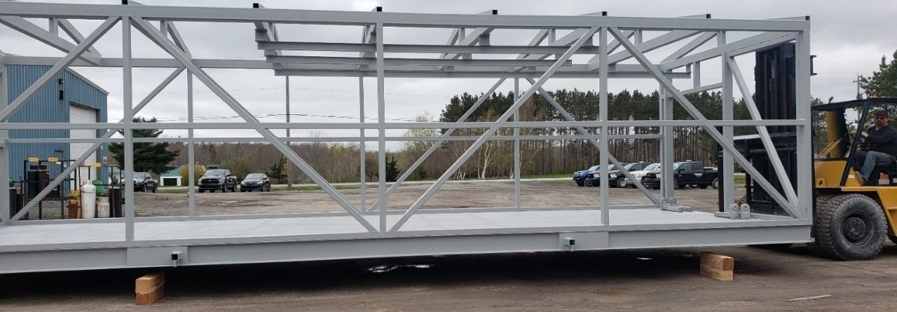 A large, gray metal frame structure is being transported by a yellow forklift outdoors, with cars, trees, and a cloudy sky in the background. The frame is resting on wooden blocks.