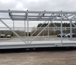 A large, gray metal frame structure is being transported by a yellow forklift outdoors, with cars, trees, and a cloudy sky in the background. The frame is resting on wooden blocks.