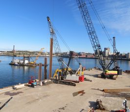 A waterfront construction site with cranes, heavy machinery, and large steel beams on a concrete dock. A barge and cranes are positioned in the water, with a city skyline visible in the background under a clear blue sky.
