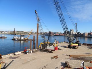 A waterfront construction site with cranes, heavy machinery, and large steel beams on a concrete dock. A barge and cranes are positioned in the water, with a city skyline visible in the background under a clear blue sky.