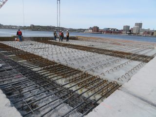 Several construction workers in safety gear stand near a concrete slab with exposed rebar at a waterfront construction site, with a city skyline and water visible in the background.