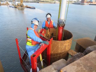 Two construction workers in safety gear and helmets stand on a red metal platform by a large cylindrical metal structure over water, with cranes and buildings visible in the background.