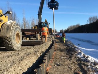 Construction workers operate heavy machinery to install vertical metal beams along a dirt road by a snow-covered area, with trees and a clear blue sky in the background.