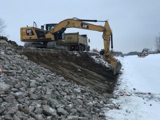 A yellow excavator is digging on a snowy, rocky embankment beside a river or canal. A dump truck is parked behind it under an overcast sky, with snow and bare trees in the background.