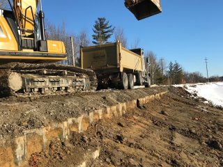 A yellow excavator loads dirt into a dump truck on a construction site beside a muddy, partially snow-covered road under a clear blue sky. Trees are visible in the background.