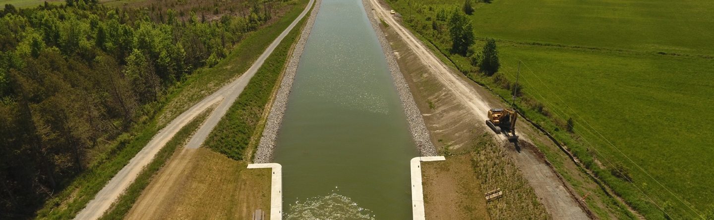 Aerial view of a wide canal with grassy banks, flanked by rural fields and trees; construction equipment is visible on the right side near the water.