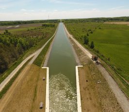 Aerial view of a wide canal with grassy banks, flanked by rural fields and trees; construction equipment is visible on the right side near the water.
