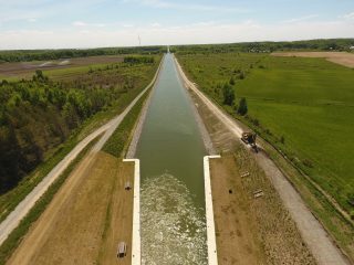 Aerial view of a wide canal with grassy banks, flanked by rural fields and trees; construction equipment is visible on the right side near the water.