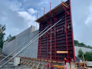 A large red metal formwork structure supports a vertical concrete wall under construction at an outdoor building site, with tools and construction materials scattered around. Trees and cloudy sky are in the background.