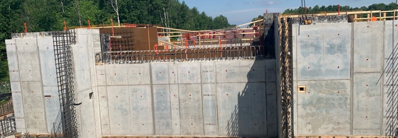 A construction site with large concrete walls, nearby metal rebar, wooden formwork, and scattered construction materials. Trees and a clear blue sky are visible in the background.