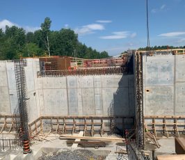 A construction site with large concrete walls, nearby metal rebar, wooden formwork, and scattered construction materials. Trees and a clear blue sky are visible in the background.