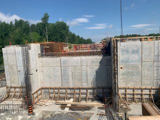 A construction site with large concrete walls, nearby metal rebar, wooden formwork, and scattered construction materials. Trees and a clear blue sky are visible in the background.