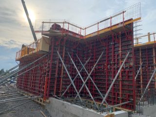 A large construction site shows red steel formwork supported by metal braces, forming a tall concrete wall under a cloudy sky. Safety railings line the top edge, and the sun is shining behind the structure.