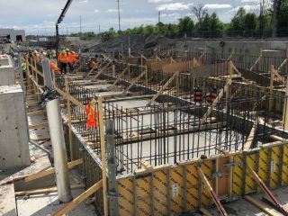 Construction workers wearing safety gear work on a large concrete foundation with exposed rebar, wooden forms, and construction equipment under a partly cloudy sky at an outdoor site.