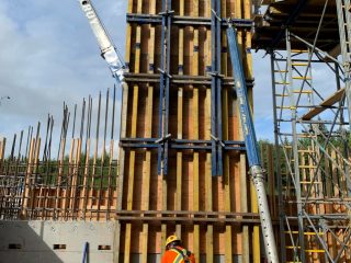 A construction worker in a safety vest and helmet stands in front of a tall wooden and metal framework at a building site, surrounded by scaffolding, rebar, and concrete walls under a partly cloudy sky.