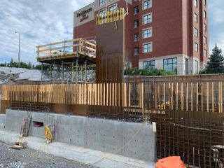 A construction site features concrete walls, vertical wooden formwork, scaffolding, and equipment, with a multi-story brick hotel building and trees in the background under a partly cloudy sky.