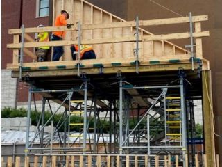 Three construction workers in safety gear stand on scaffolding, working on a wooden structure attached to a building under construction. The background shows a partially completed building and various materials.