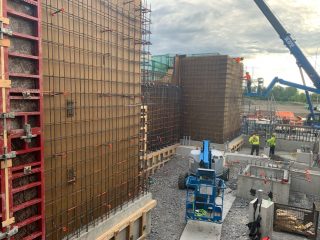 Construction site with workers, steel reinforcement bars, wooden and metal formwork, a blue lift, concrete walls, and a crane against a cloudy sky. Building materials and equipment are scattered around the area.