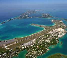 Aerial view of an island with turquoise water, an airport runway, and scattered buildings; the land extends into the distance, surrounded by the sea under a clear sky.