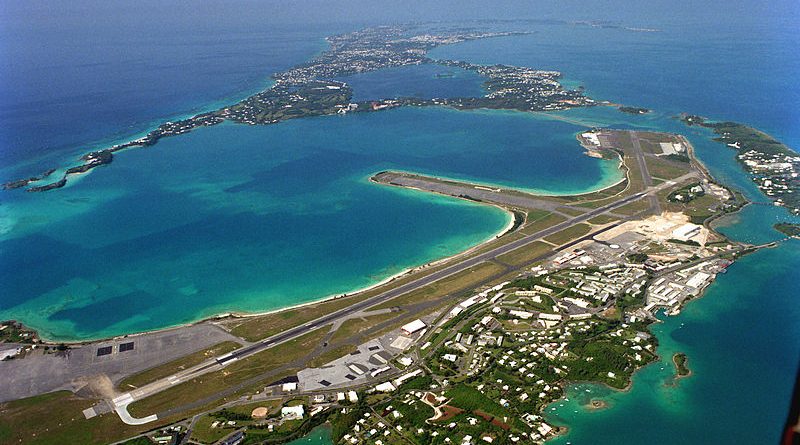 Aerial view of an island with turquoise water, an airport runway, and scattered buildings; the land extends into the distance, surrounded by the sea under a clear sky.