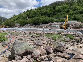 A yellow excavator works beside a partially built stone wall in a rocky, forested area with scattered boulders and a cloudy sky overhead. Construction materials and pipes are visible on the ground.