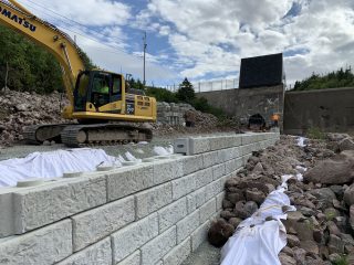 A yellow excavator sits on gravel near a newly constructed stone retaining wall, with two workers and equipment further along the wall. The scene includes rocks, white fabric, and a partly cloudy sky.