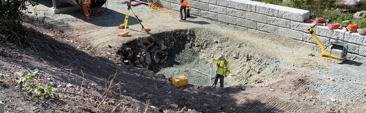 Several construction workers are installing a large metal culvert beside a gravel road surrounded by trees and rocks, with safety barriers and equipment visible nearby.