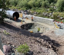 Several construction workers are installing a large metal culvert beside a gravel road surrounded by trees and rocks, with safety barriers and equipment visible nearby.