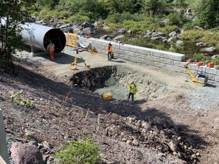 Several construction workers are installing a large metal culvert beside a gravel road surrounded by trees and rocks, with safety barriers and equipment visible nearby.